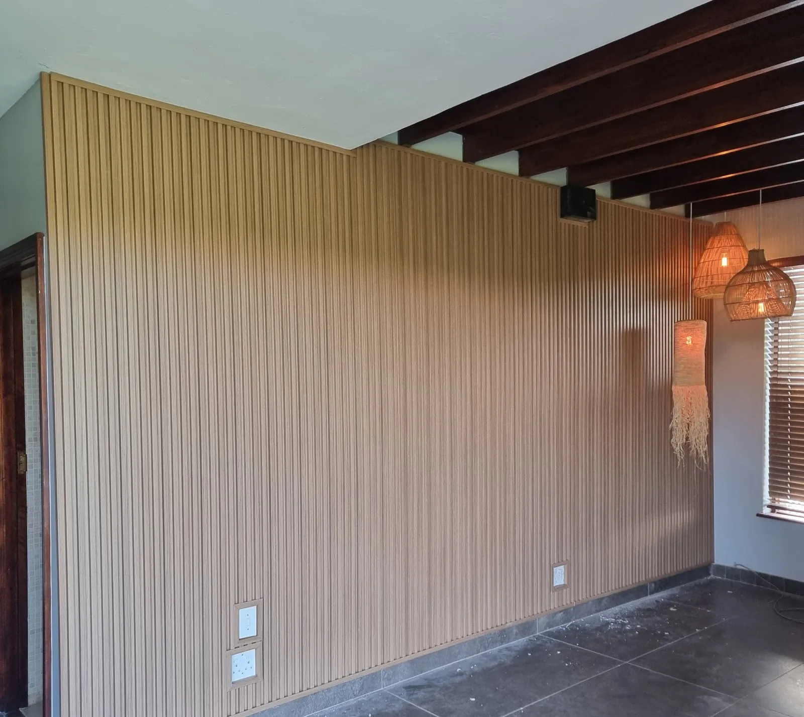 A wall clad floor to ceiling in vertical slatted panels, beneath dark timber ceiling beams